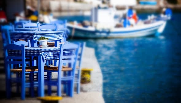 Set of blue wooden chairs and a table with a checkered tablecloth near a body of water. - Olive Oil Times