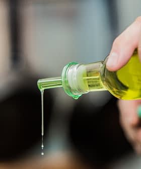 A person pouring olive oil from a glass bottle with a green cap, with oil dripping from the spout. - Olive Oil Times