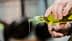 A person pouring olive oil from a glass bottle with a green cap, with oil dripping from the spout. - Olive Oil Times