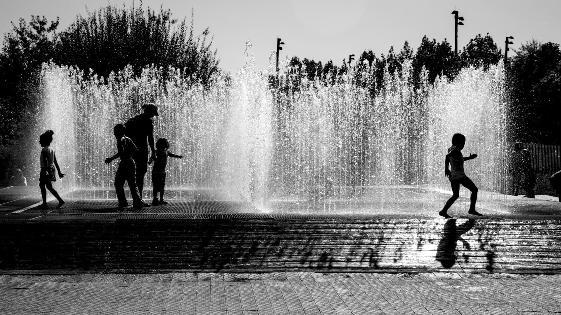 Silhouettes of children playing in a water fountain with water spraying in the background. - Olive Oil Times