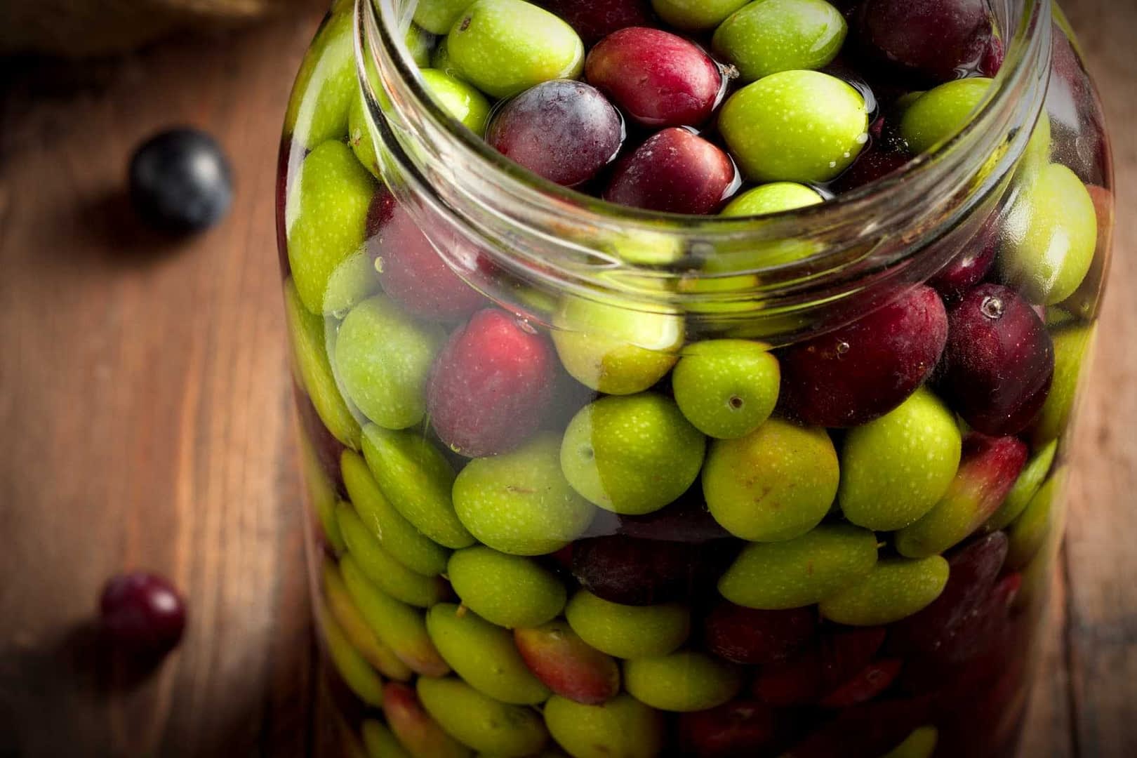 Glass jar filled with a mix of green and black olives on a wooden surface. - Olive Oil Times
