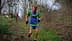 Two trail runners participating in a race on a wooded path, one wearing a green and blue outfit with a race number. - Olive Oil Times
