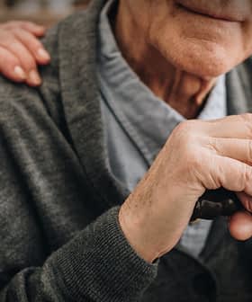 Close-up of an elderly man's hands resting on a cane, with a hand on his shoulder. - Olive Oil Times