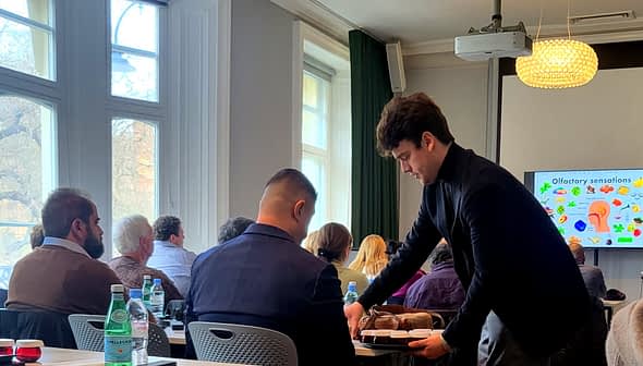 A young man serving food to a seated participant during a presentation on olfactory sensations. - Olive Oil Times