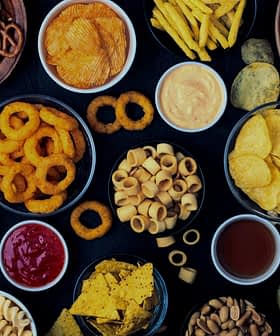 An assortment of snack foods including chips, pretzels, and popcorn arranged in bowls on a dark surface. - Olive Oil Times