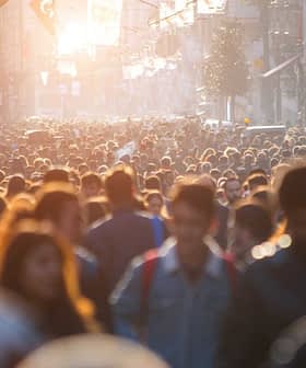 A large crowd of people walking in a busy urban street during sunset with sunlight filtering through. - Olive Oil Times