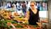 A woman wearing sunglasses examines produce at a farmers market with various fruits and vegetables displayed. - Olive Oil Times