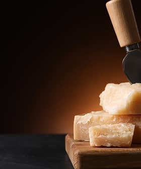 Block of Parmesan cheese with a cheese knife inserted into it, resting on a wooden cutting board. - Olive Oil Times