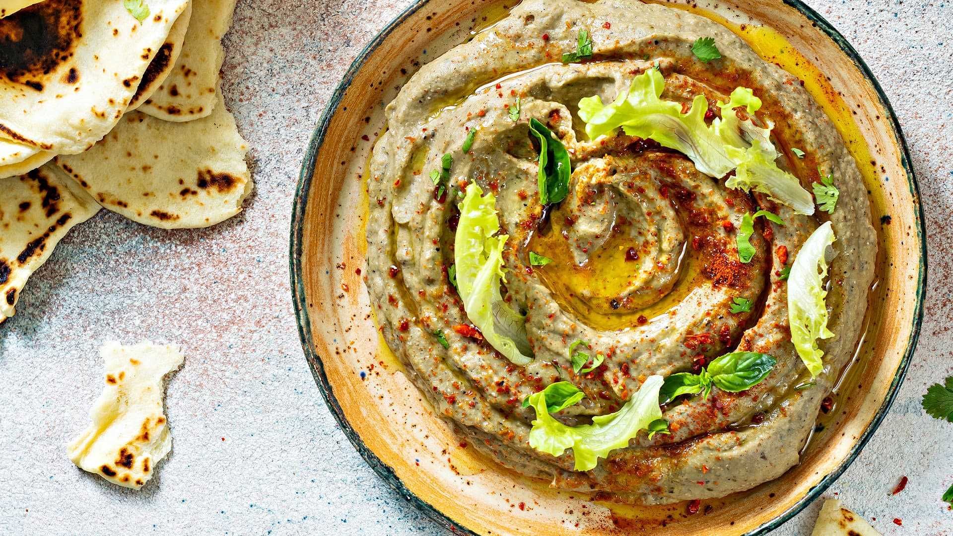 Bowl of hummus topped with olive oil, herbs, and spices, served with flatbread on the side. - Olive Oil Times