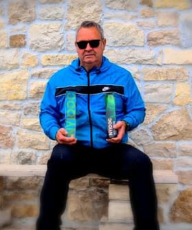 Man wearing sunglasses and a blue jacket holding two bottles of olive oil while seated on a stone bench. - Olive Oil Times