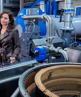 A woman in a brown leather jacket conversing with a man in a factory filled with machinery and equipment. - Olive Oil Times