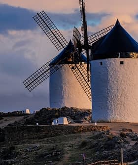 Two traditional windmills with black blades and white stone structures situated on a hilltop. - Olive Oil Times
