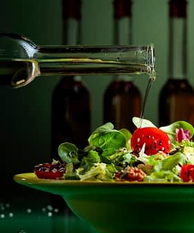 A hand pouring olive oil from a glass bottle onto a salad in a green bowl with cherry tomatoes and greens. - Olive Oil Times