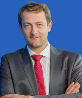 Man wearing a gray suit and a red tie, standing with arms crossed against a blue background. - Olive Oil Times