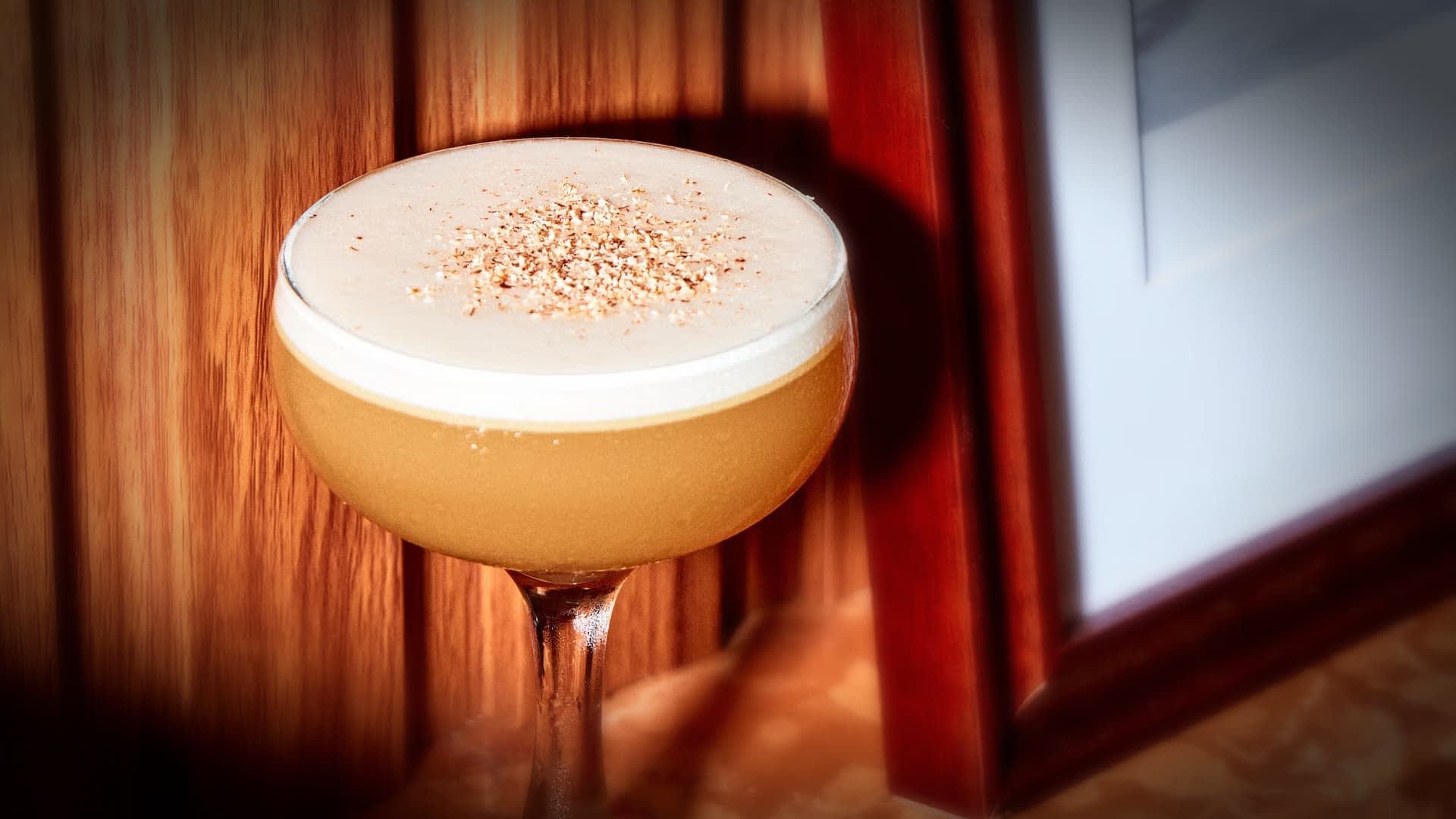 A cocktail in a coupe glass with a frothy top, garnished with grated nutmeg, is placed on a wooden surface next to a framed picture. The background is a wooden panel wall.