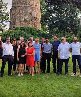 A group of individuals posing together for a photo in an outdoor area with greenery and a stone structure. - Olive Oil Times
