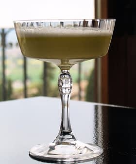 A crystal glass filled with a light green cocktail placed on a reflective surface. - Olive Oil Times