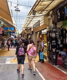 Two people walking through a market street lined with shops and stalls selling various items. - Olive Oil Times