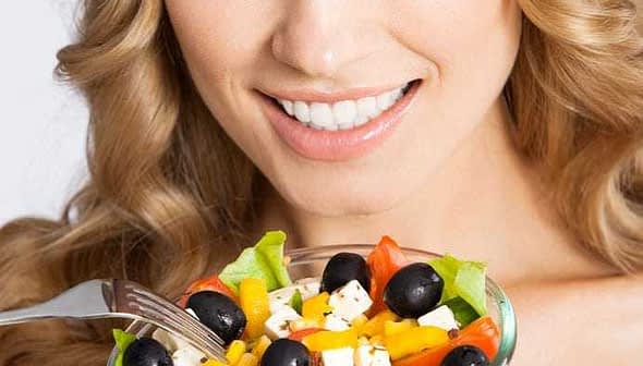 Woman with long blonde hair smiling while holding a bowl of colorful salad with vegetables and olives. - Olive Oil Times
