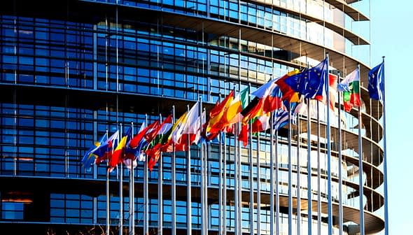 Multiple flags of European countries displayed in front of a modern glass building. - Olive Oil Times