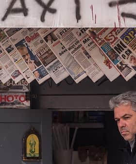 A man standing in front of a newsstand displaying various newspapers and magazines. - Olive Oil Times