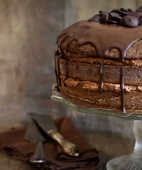 Chocolate layer cake topped with chocolate ganache and chocolate chips on a glass cake stand. - Olive Oil Times