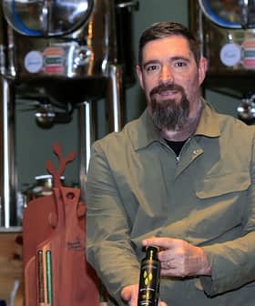 Man with a beard holding a bottle of olive oil in a production facility with metal containers in the background. - Olive Oil Times
