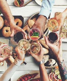 Group of people toasting with glasses over a table filled with various foods and drinks. - Olive Oil Times