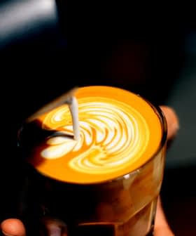 A glass of latte with intricate milk foam art on top, held in a hand against a dark background. - Olive Oil Times