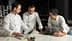 Three chefs in white jackets examining a cookbook while discussing culinary presentations. - Olive Oil Times