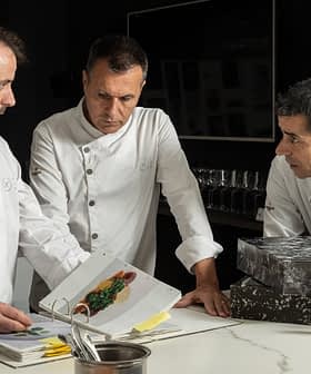 Three chefs in white jackets examining a cookbook while discussing culinary presentations. - Olive Oil Times