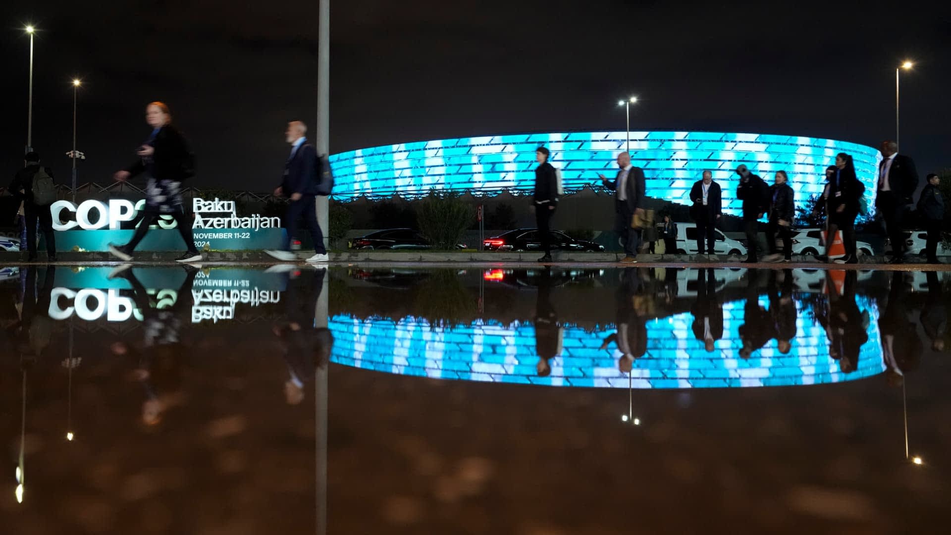 Group of people walking past a modern building with illuminated blue facade at night, reflected in a puddle. - Olive Oil Times