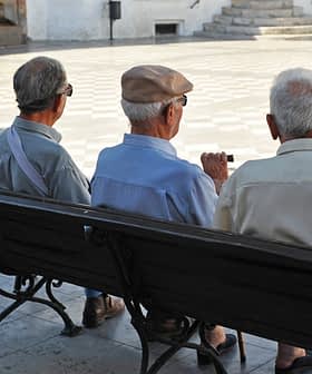 Three elderly men sitting on a bench, viewed from behind, in a public square. - Olive Oil Times