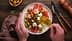 Hands pouring olive oil over a salad with tomatoes, cucumbers, olives, and feta cheese on a plate. - Olive Oil Times