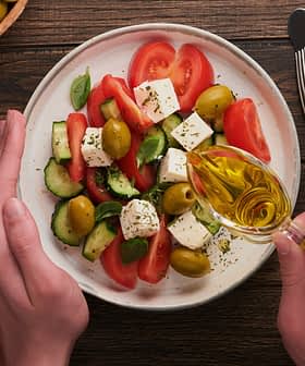 Hands pouring olive oil over a salad with tomatoes, cucumbers, olives, and feta cheese on a plate. - Olive Oil Times