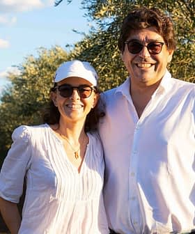 A man and woman smiling while standing in an olive grove under a clear sky. - Olive Oil Times