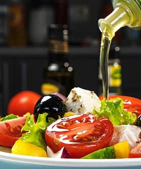 A bowl of fresh salad being drizzled with olive oil, featuring tomatoes, lettuce, and olives. - Olive Oil Times