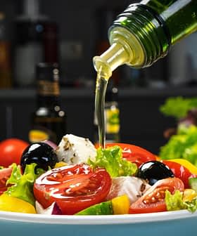 Olive oil being poured over a fresh salad with tomatoes, lettuce, and olives in a bowl. - Olive Oil Times