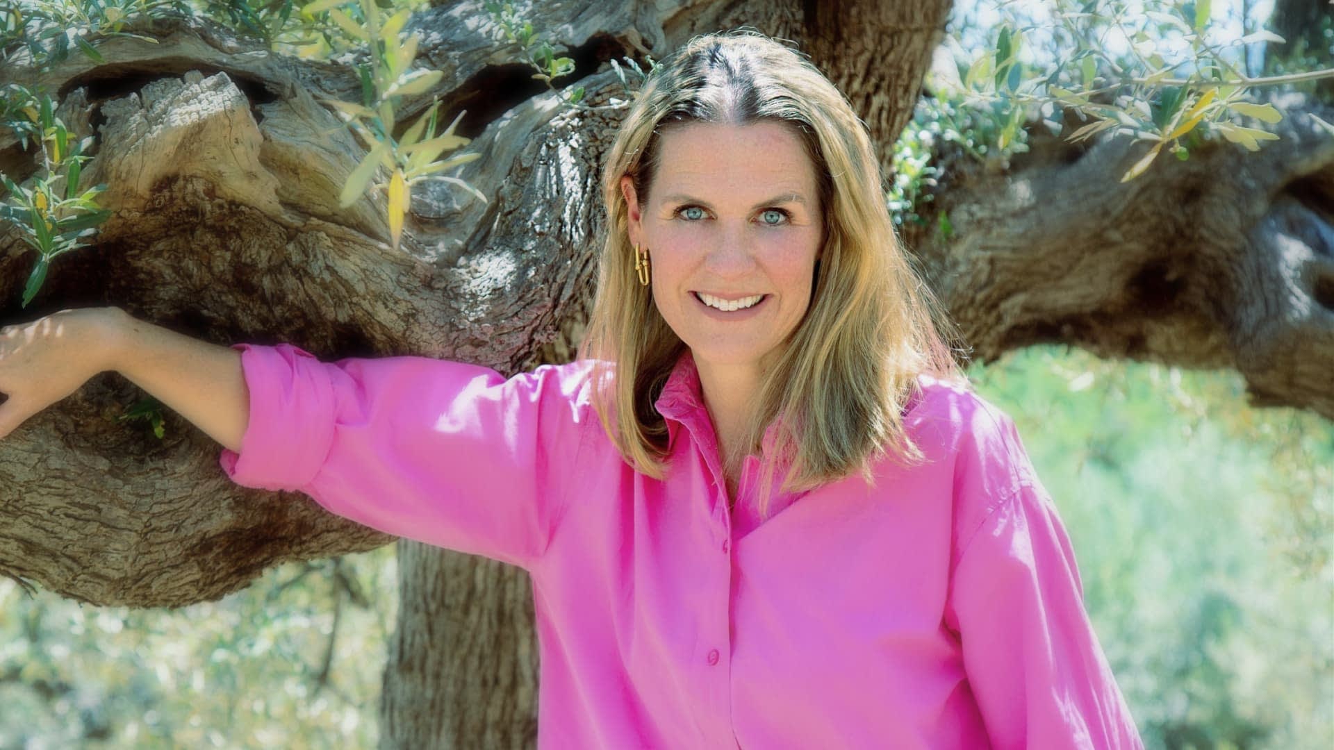 Woman with long hair wearing a pink shirt, leaning against an olive tree with a smile. - Olive Oil Times