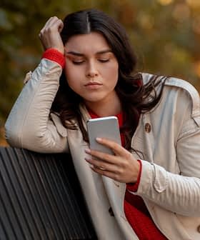 A woman with long dark hair wearing a light coat and red sweater, looking at her smartphone while sitting on a bench. - Olive Oil Times