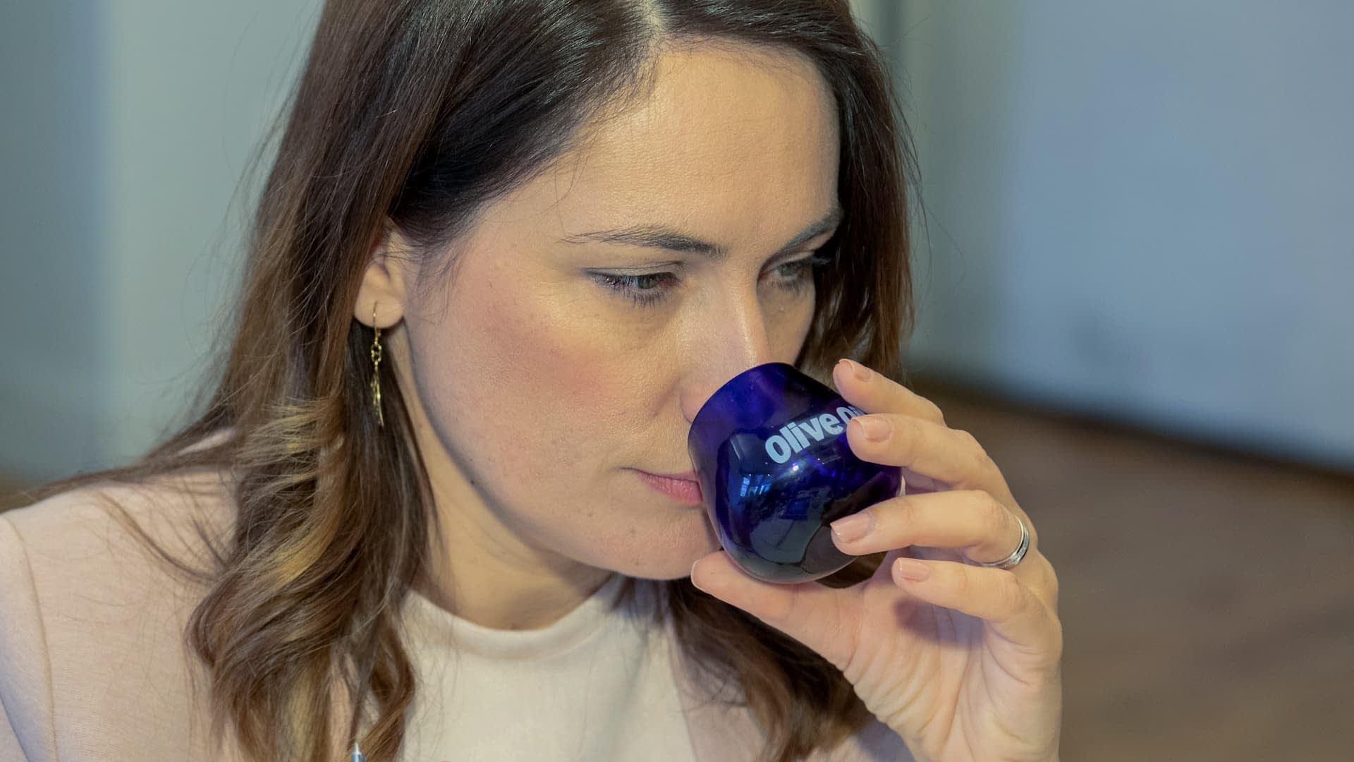 Woman with brown hair holding a blue tasting glass labeled 'Olive Oil' close to her face. - Olive Oil Times