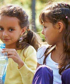 Two young girls sitting outdoors, one holding a plastic water bottle and looking at the other. - Olive Oil Times