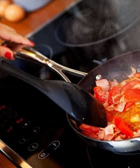 Person stirring a mixture of chopped tomatoes and bacon in a frying pan on a stovetop. - Olive Oil Times
