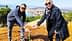 Two individuals bending down to plant a small tree in a rural area with a view of distant hills and water. - Olive Oil Times