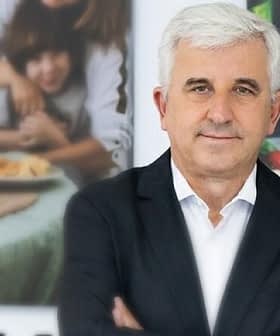 A man with white hair wearing a black suit, standing with arms crossed in front of a blurred background. - Olive Oil Times