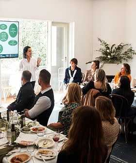 A speaker presenting information on a screen to an audience seated at a long table during a meeting. - Olive Oil Times