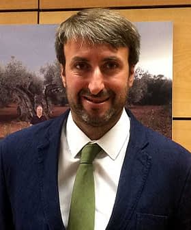 Man wearing a dark suit and a green tie, standing in front of a background featuring olive trees. - Olive Oil Times