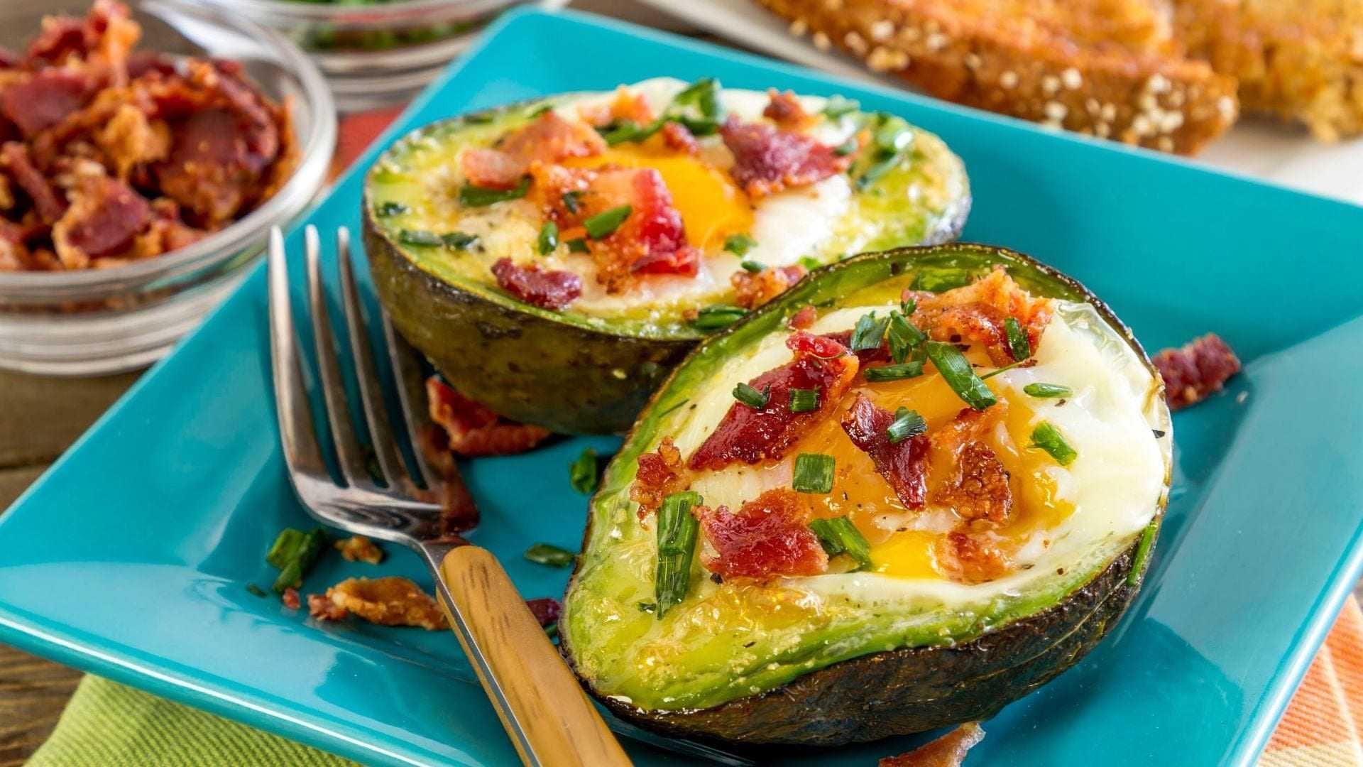 Baked avocado halves filled with egg, topped with bacon bits and chives on a blue plate. - Olive Oil Times