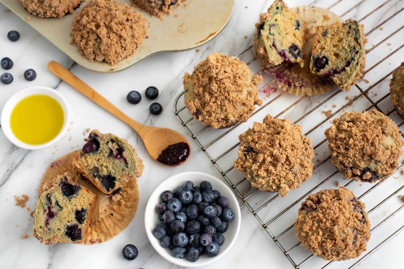 Freshly baked blueberry muffins with a crumb topping on a cooling rack and a plate of blueberries. - Olive Oil Times