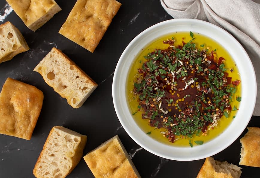 Bowl of olive oil mixed with herbs and spices surrounded by pieces of bread on a dark surface. - Olive Oil Times